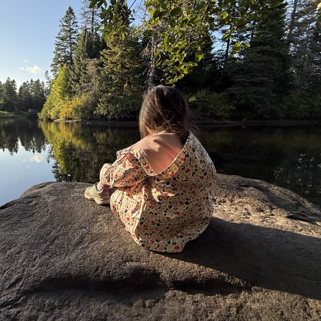 Sa plus jeune, de dos, assise sur un rocher devant la rivière