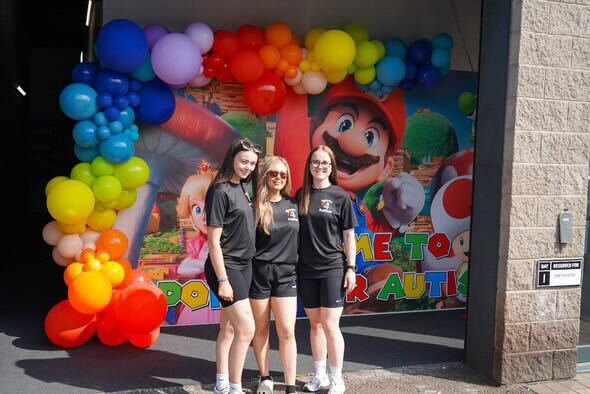 Three coaches in front of a Mario-themed welcome backdrop and balloon arch at the SFA venue