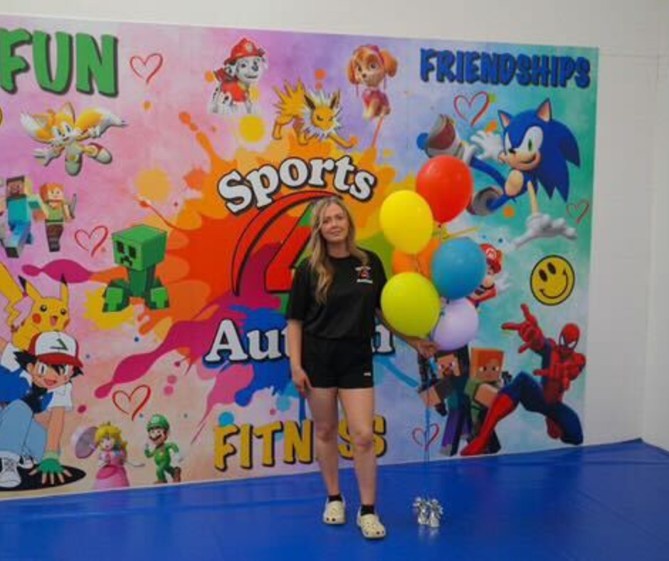 Stacey holding a bunch of balloons in front of the colourful Sports 4 Autism mural that reads Fun, Fitness, Friendships