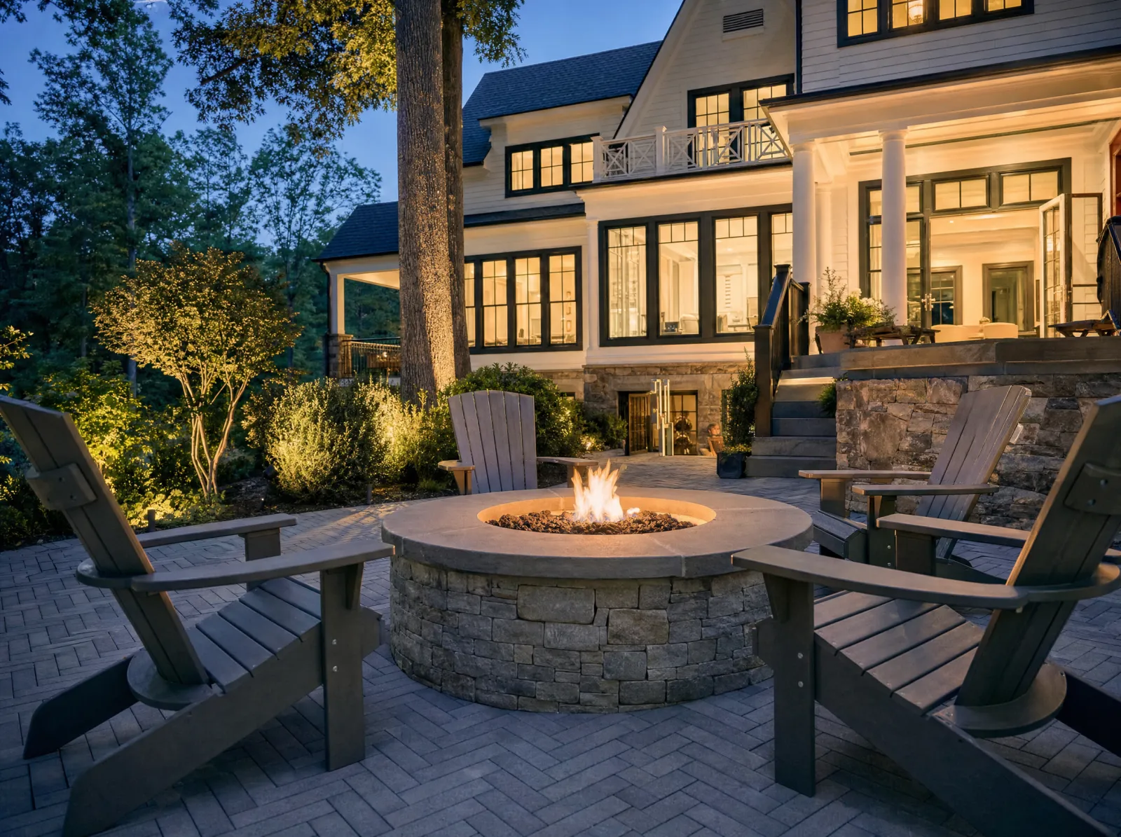 Design concept: Round gas fire pit in ledgestone veneer with a bluestone cap, flames lit, four Adirondack chairs, paver patio, blue hour.