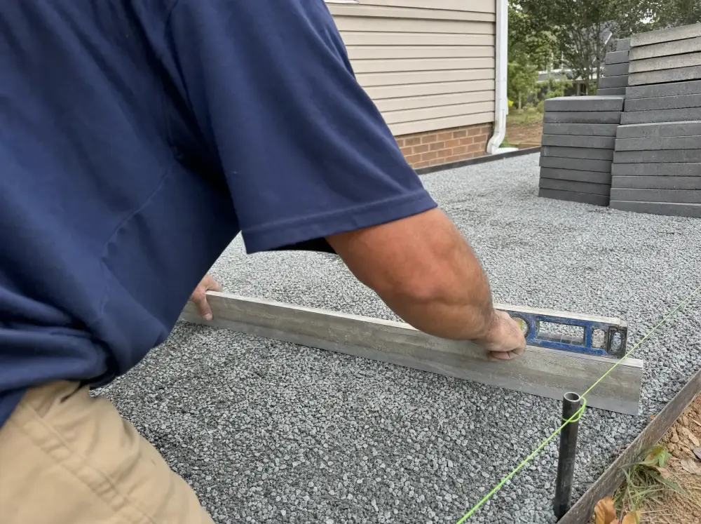 Illustration: a straightedge and torpedo level laid across a compacted crushed-stone base, string line set along the edge, pavers stacked to one side.