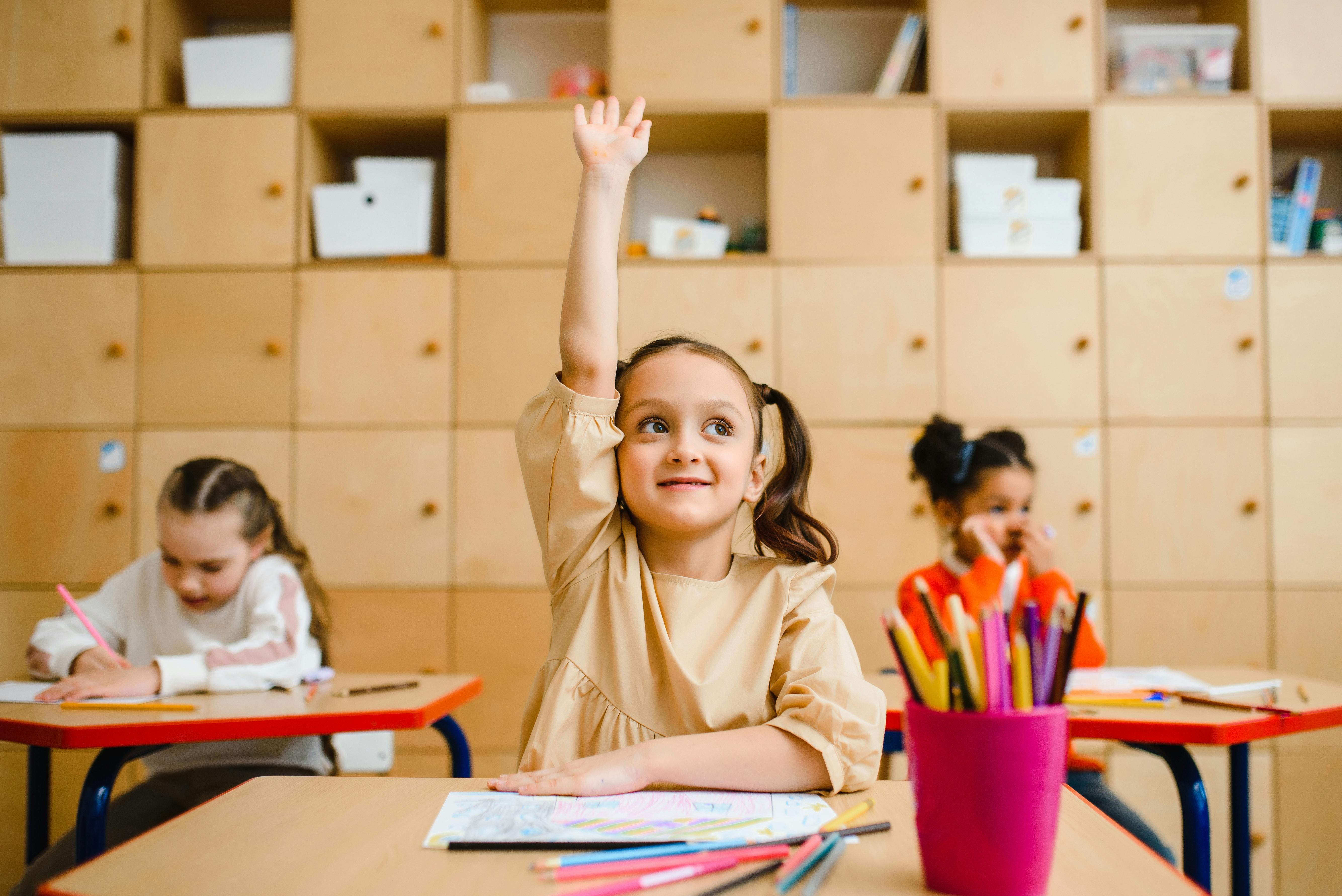 An image of a young girl raising her hand in a classroom An image of a young girl raising her hand in a classroom