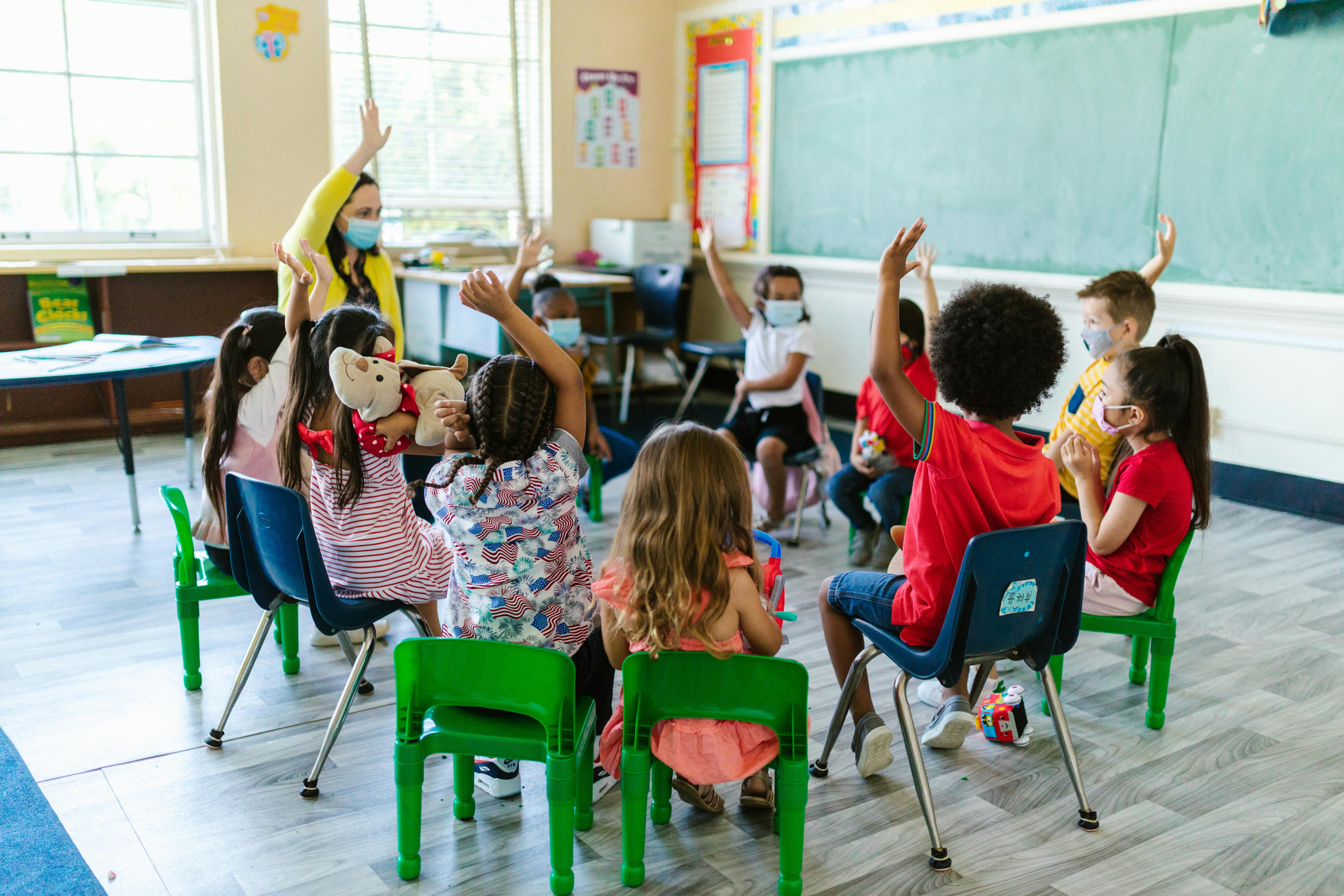 An image of children sitting in a circle in a classroom, raising their hands An image of children sitting in a circle in a classroom, raising their hands