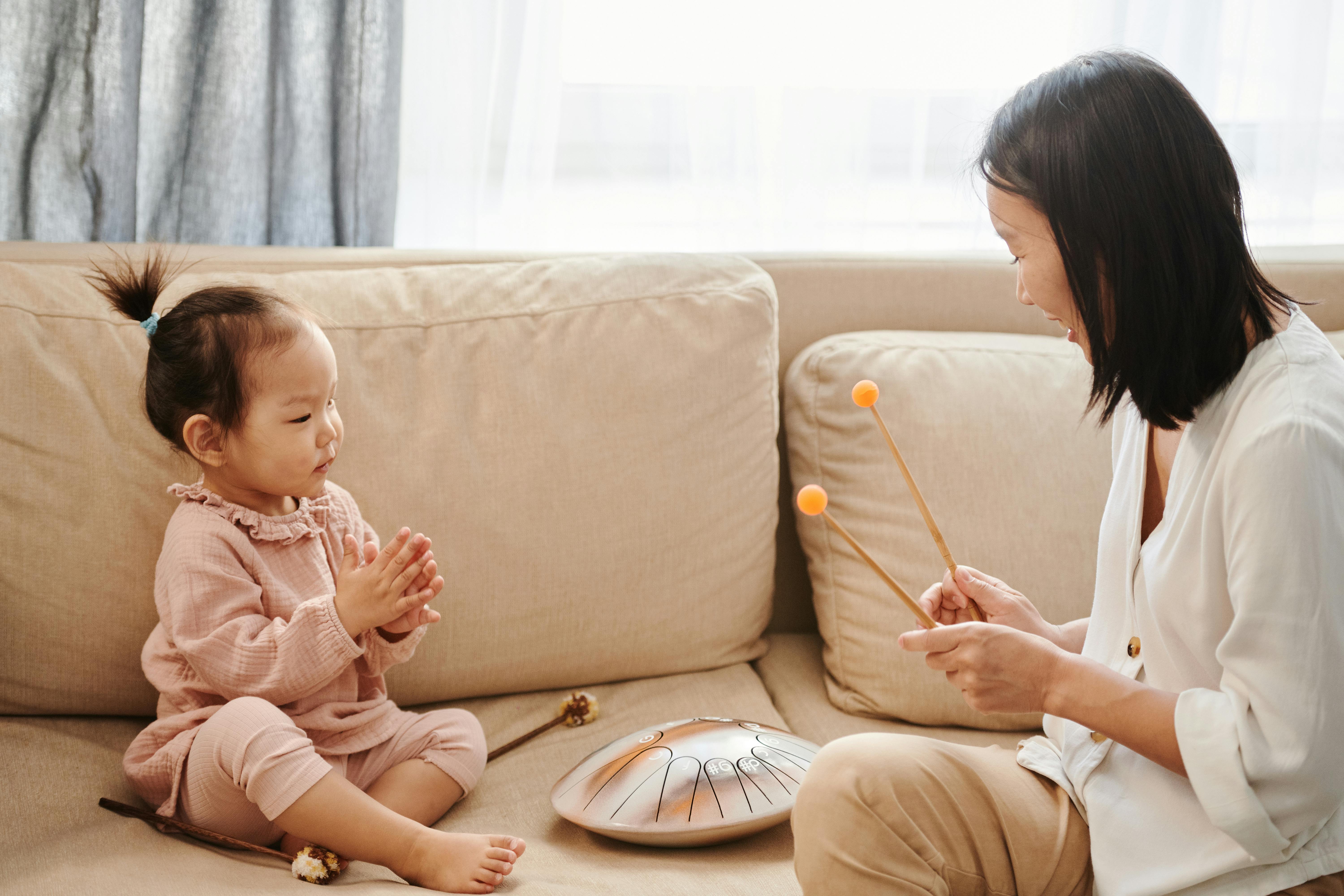  A mother and toddler sitting on a couch playing with a hand drum together.