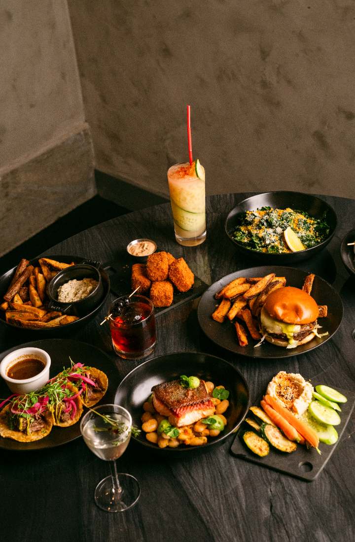 A table of Kaya dishes: tacos, wings, burger, salmon, and cocktails