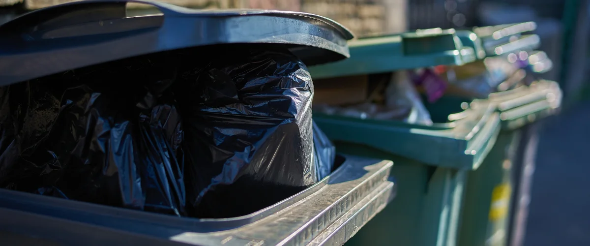 A close-up view of an overflowing black garbage bin with a slightly open lid, filled with black trash bags. A close-up view of an overflowing black garbage bin with a slightly open lid, filled with black trash bags.