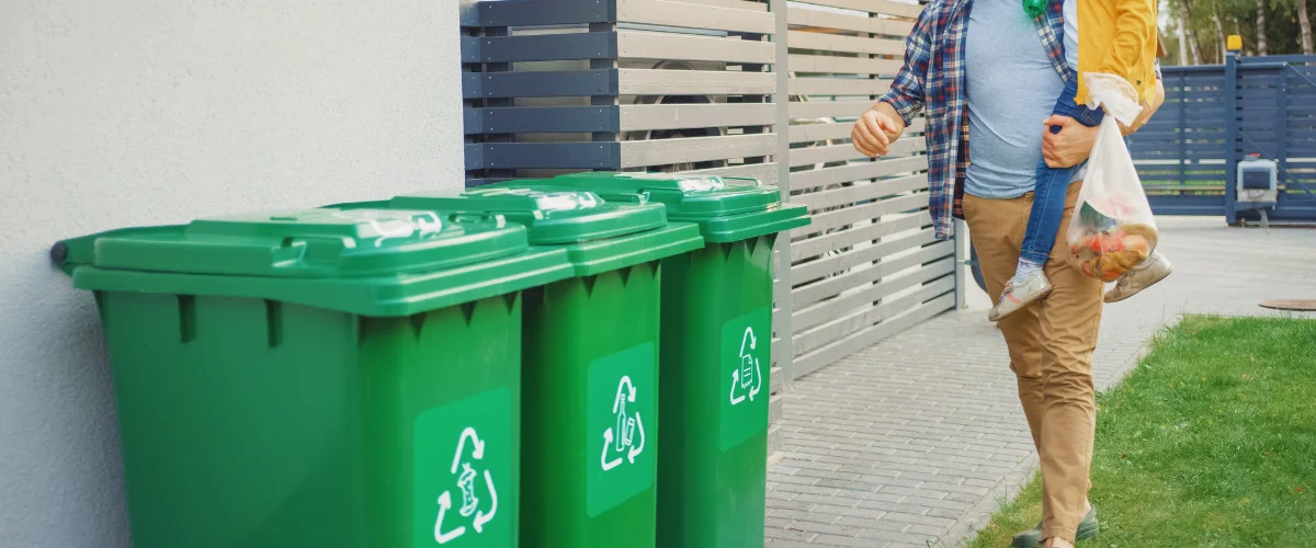 A man carries a young child and a bag of groceries past a row of green recycling bins with white symbols. A man carries a young child and a bag of groceries past a row of green recycling bins with white symbols.