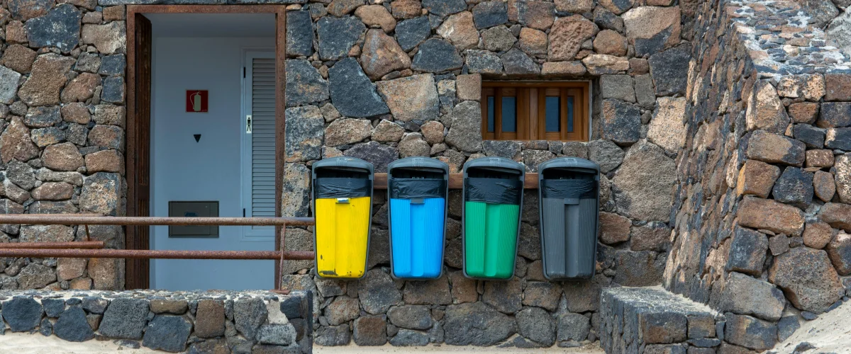 Four colorful recycling bins, yellow, blue, green, and gray, hanging on a textured stone wall next to an open doorway. Four colorful recycling bins, yellow, blue, green, and gray, hanging on a textured stone wall next to an open doorway.