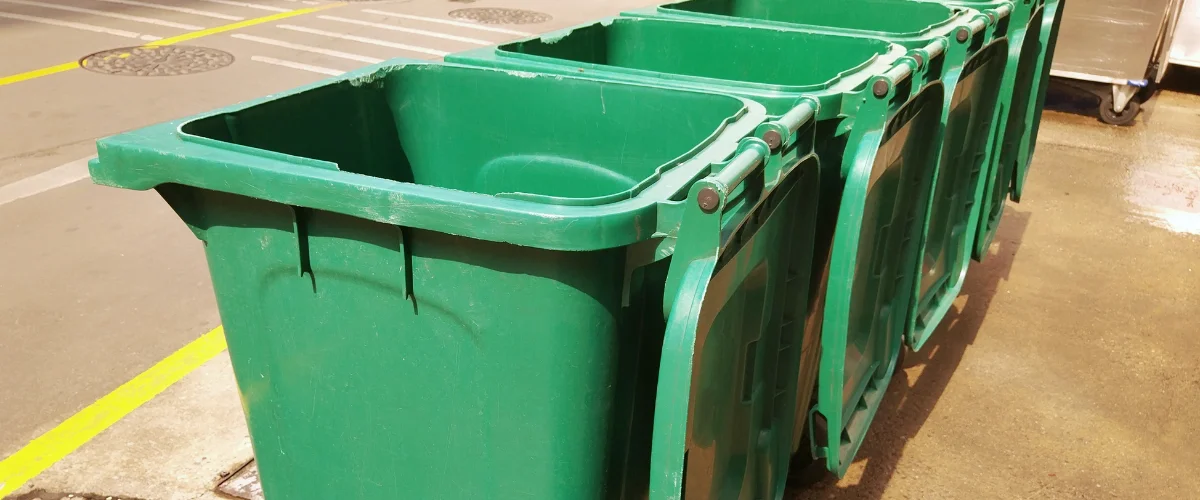A row of four green recycling bins with open lids stand along a yellow-lined pavement on a sunny day. A row of four green recycling bins with open lids stand along a yellow-lined pavement on a sunny day.