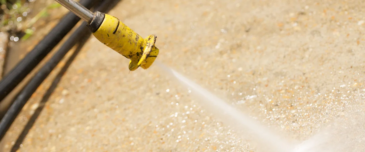 A close-up of a yellow pressure washer nozzle cleaning a speckled concrete surface with a stream of water. A close-up of a yellow pressure washer nozzle cleaning a speckled concrete surface with a stream of water.