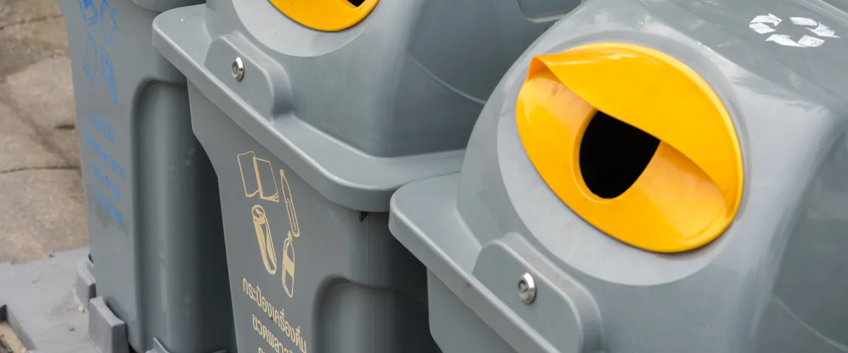 A close-up view of two grey outdoor recycling bins with bright yellow, crescent-shaped disposal openings. A close-up view of two grey outdoor recycling bins with bright yellow, crescent-shaped disposal openings.