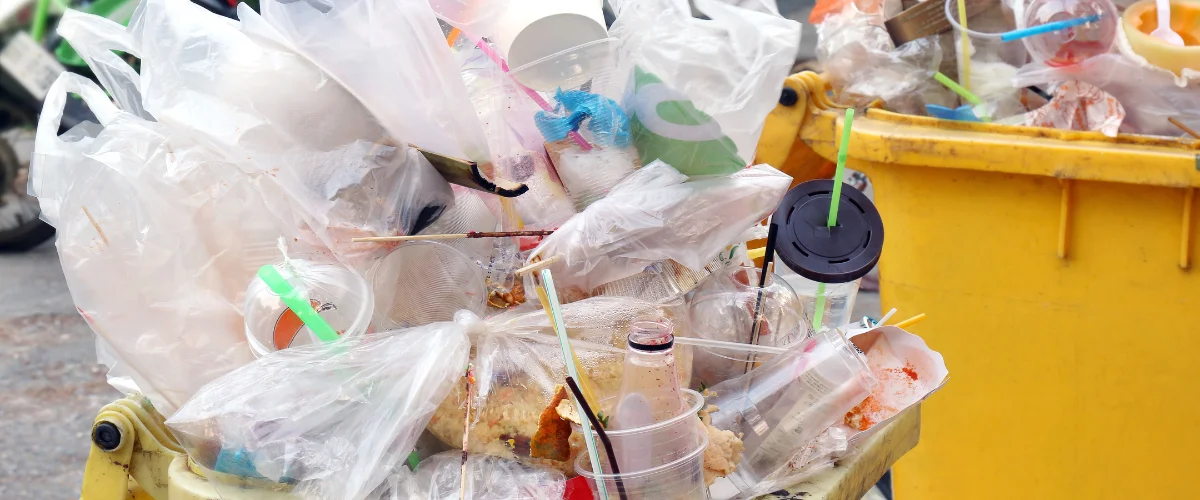 A bright yellow outdoor trash bin overflowing with plastic bags, cups, straws, and other disposable waste. A bright yellow outdoor trash bin overflowing with plastic bags, cups, straws, and other disposable waste.