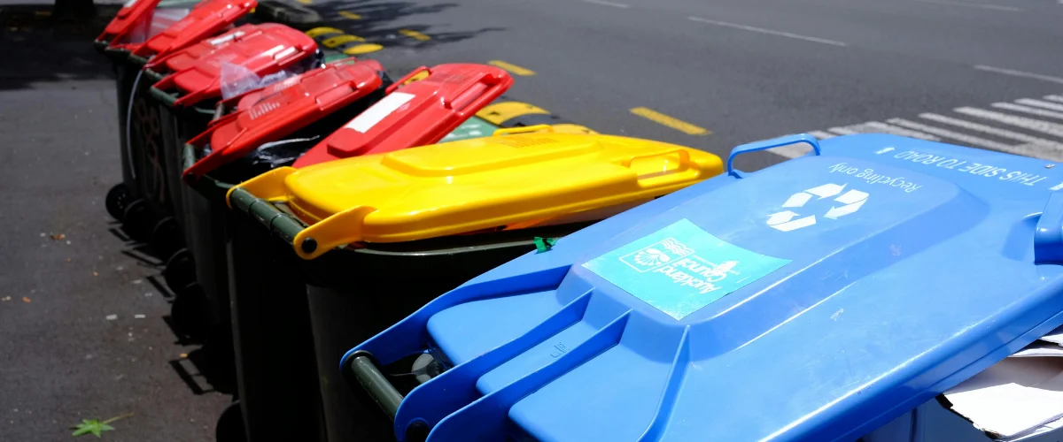 A long row of green garbage bins with colorful lids, some green, some yellow, and some red, lined up outside on a sunny day. A long row of green garbage bins with colorful lids, some green, some yellow, and some red, lined up outside on a sunny day.