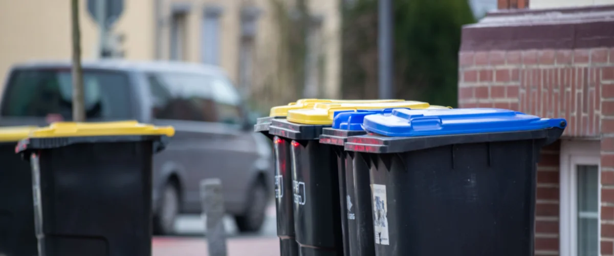 A row of several blue and black plastic garbage bins with one black bin lid open, on a suburban street. A row of several blue and black plastic garbage bins with one black bin lid open, on a suburban street.