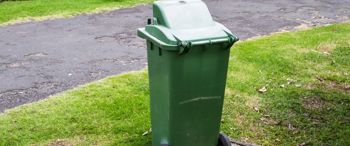 A tall, green garbage can with a closed lid on small wheels sits on lush green grass next to a worn asphalt driveway. A tall, green garbage can with a closed lid on small wheels sits on lush green grass next to a worn asphalt driveway.