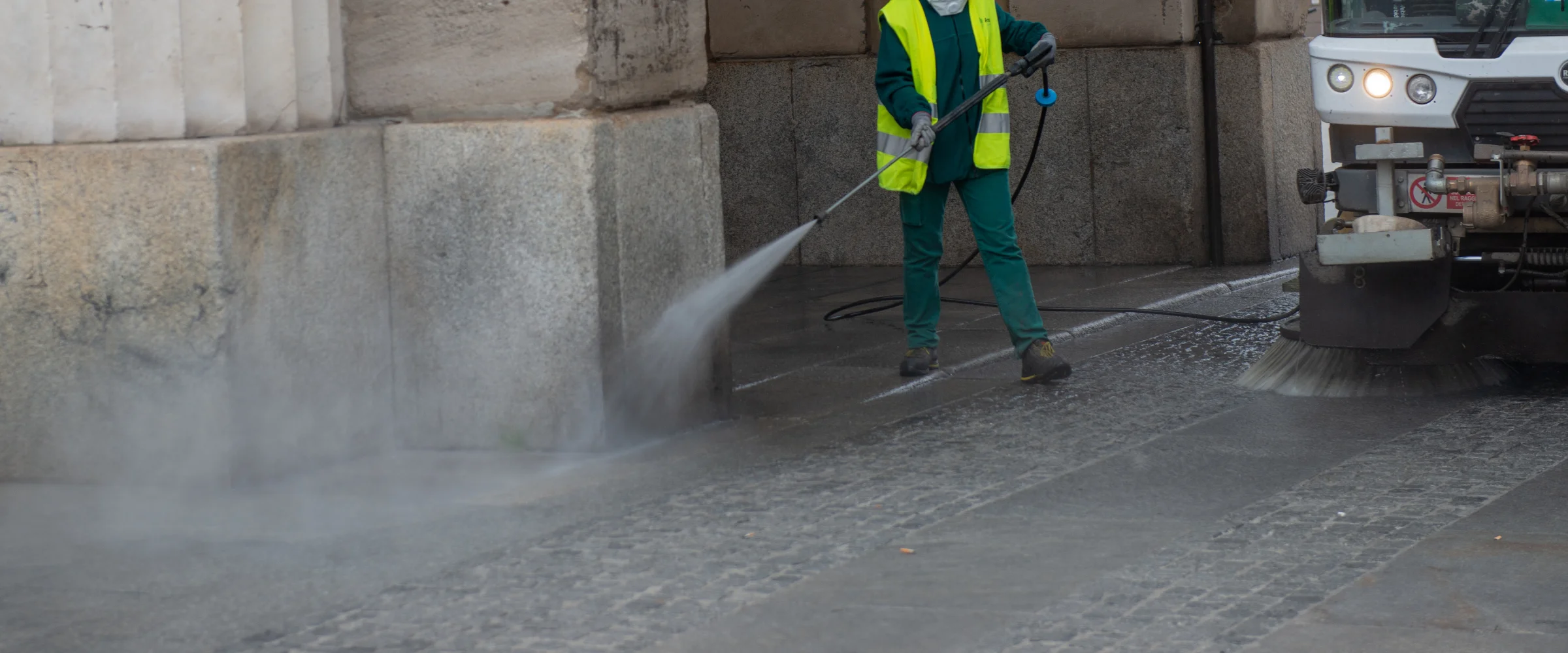 A worker in a high-vis vest power washes a cobbled street next to a street cleaning vehicle with a rotating brush. A worker in a high-vis vest power washes a cobbled street next to a street cleaning vehicle with a rotating brush.