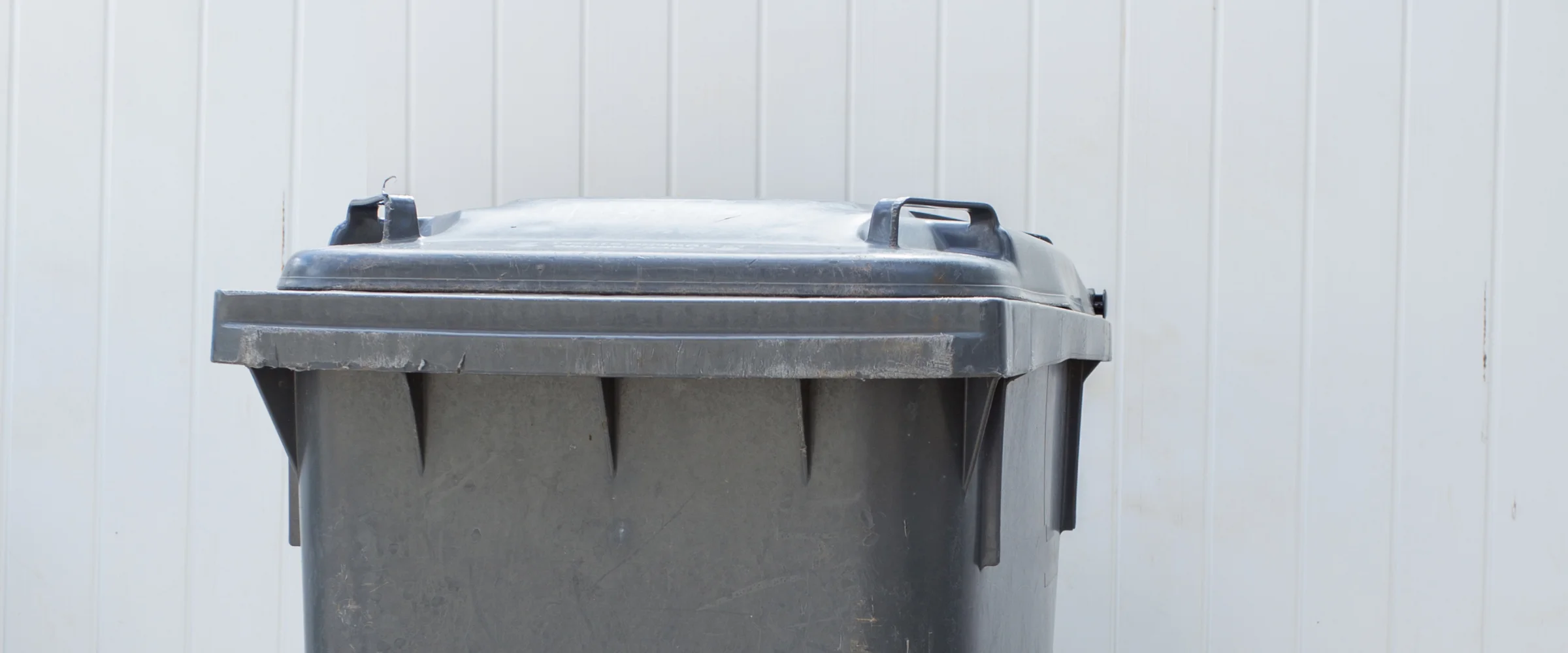 A weathered dark grey plastic trash can with a closed lid stands against a light grey slatted fence.