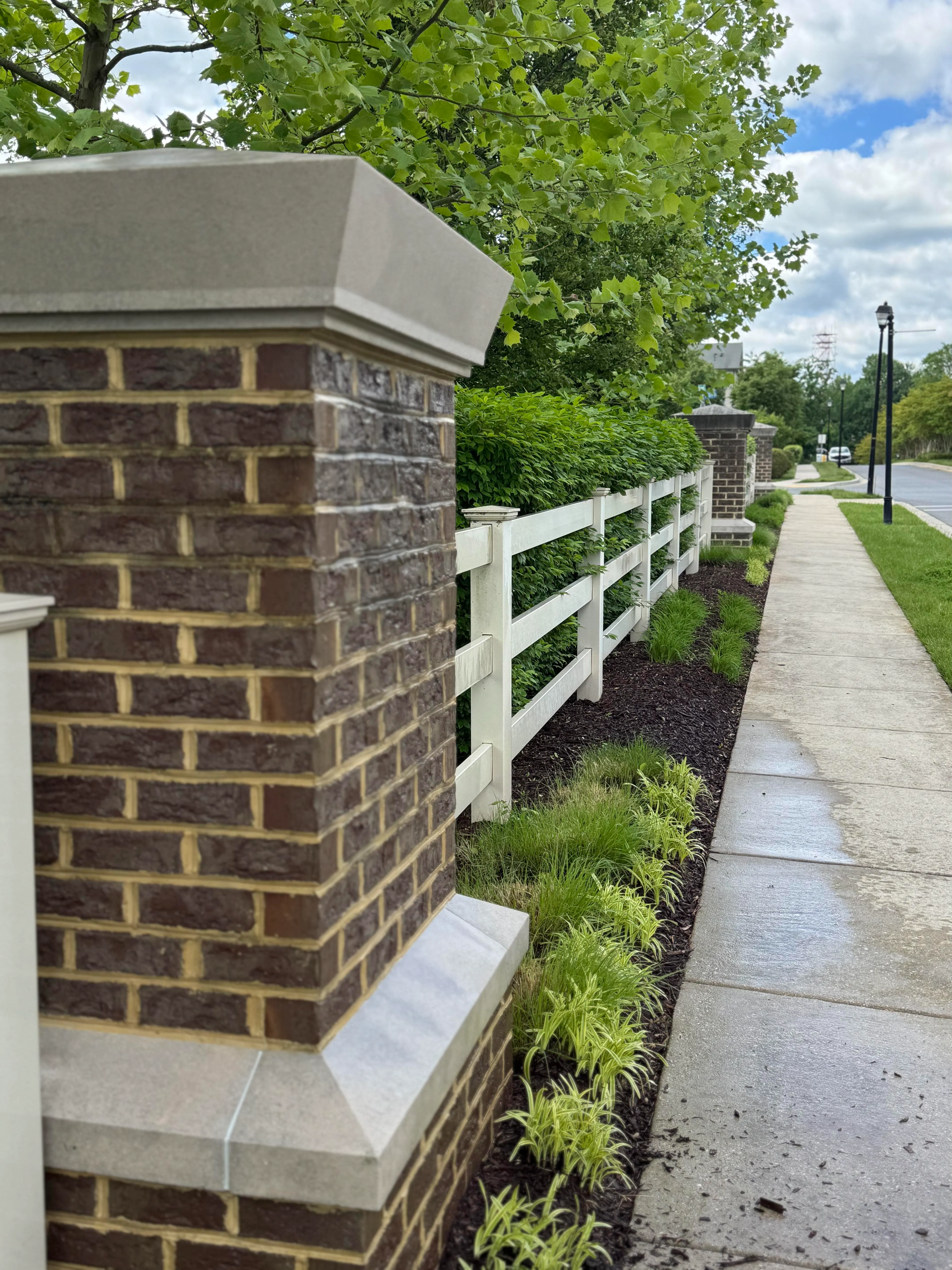 A close-up of a brick pillar and white fence next to a sidewalk with green landscaping and trees.