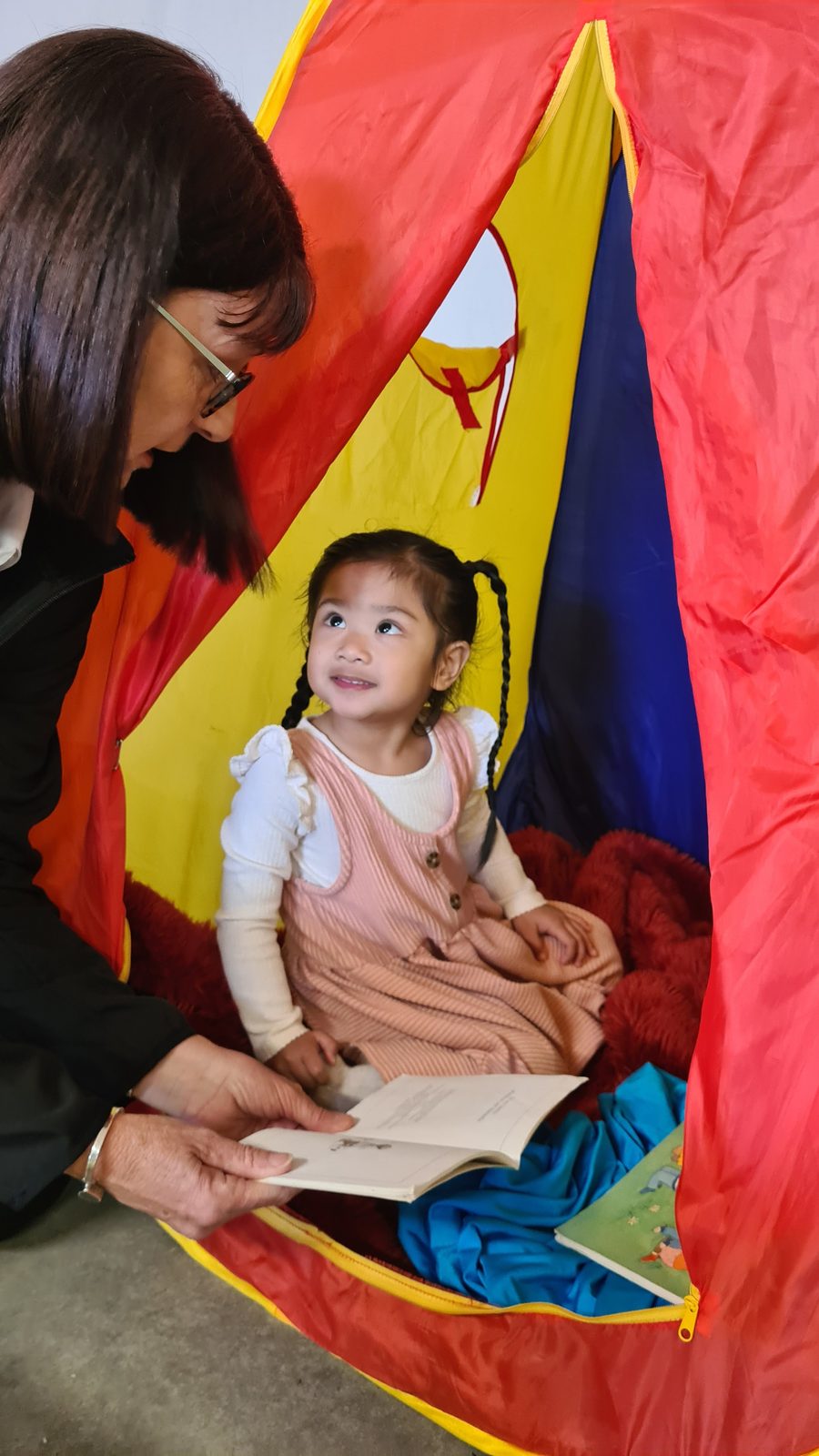 A young child sits in a play tent looking up at an adult who is holding a book