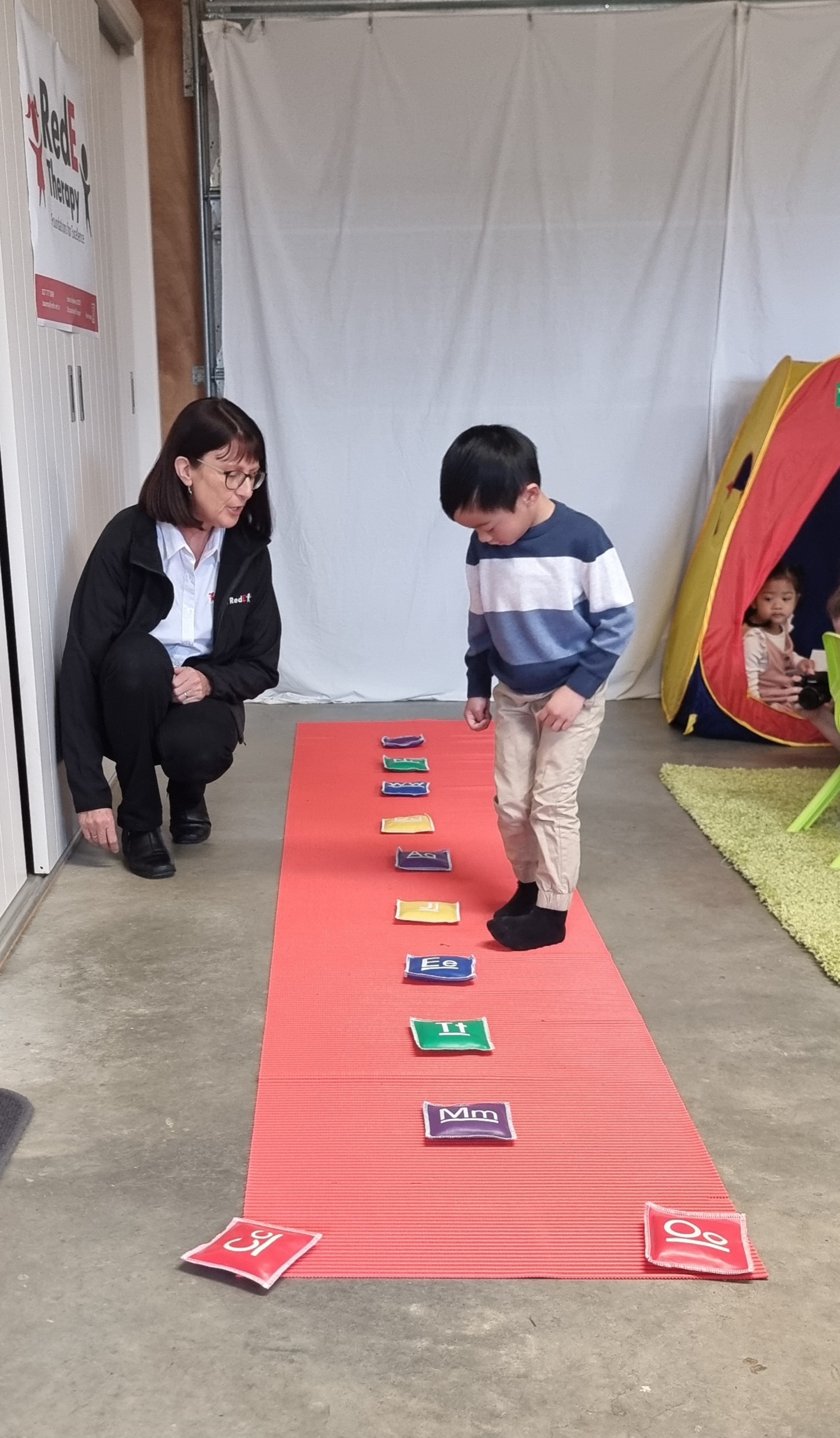 Joanna Hawkins crouching beside a boy as he looks down at letter beanbags on a red mat