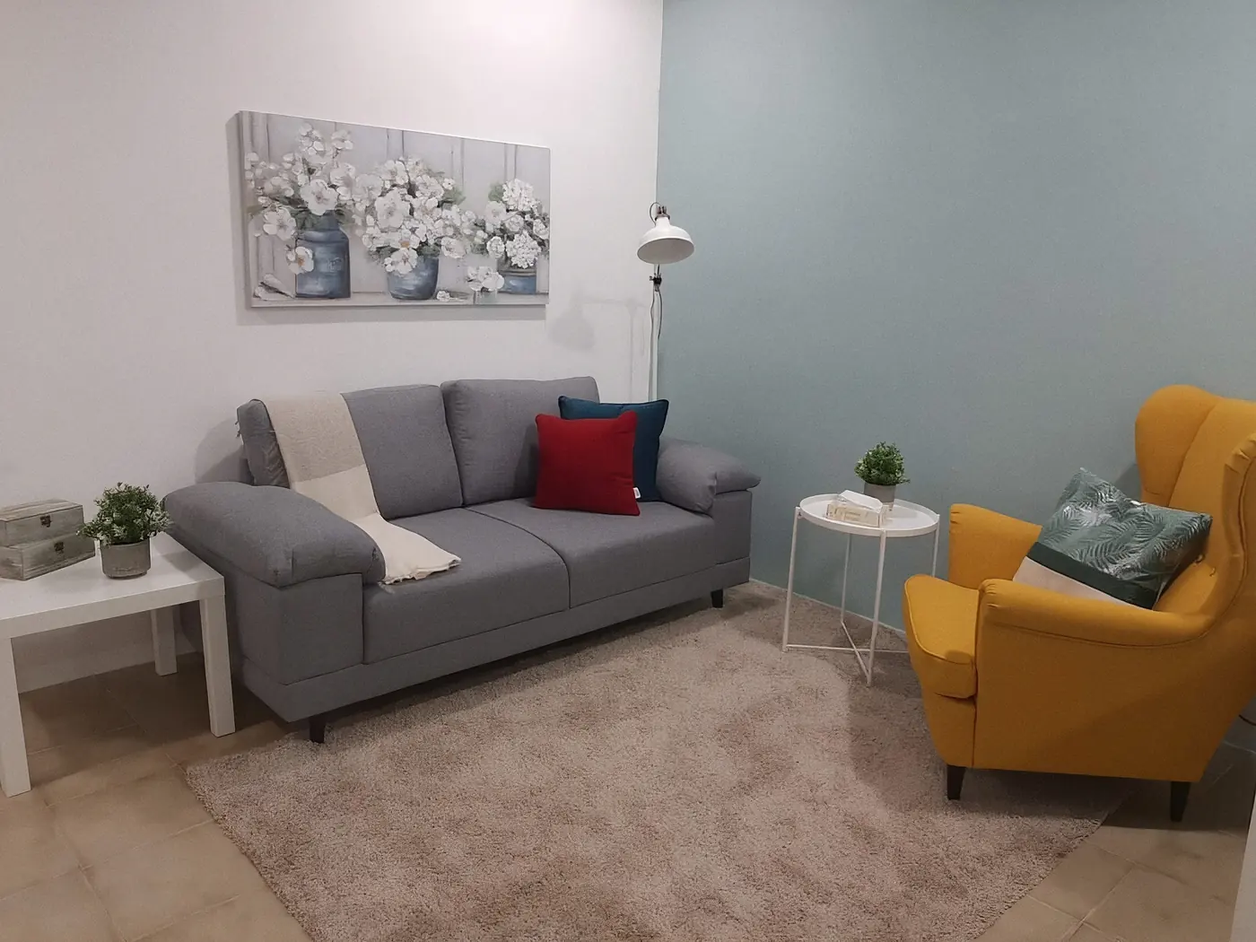 Sala de consulta com sofá cinzento, poltrona amarela, tapete claro e quadro de flores brancas