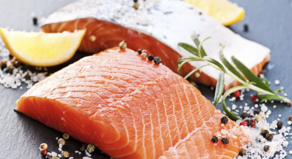 Two raw salmon fillets on a dark slate surface, garnished with coarse salt, mixed peppercorns, fresh rosemary, and lemon wedges.