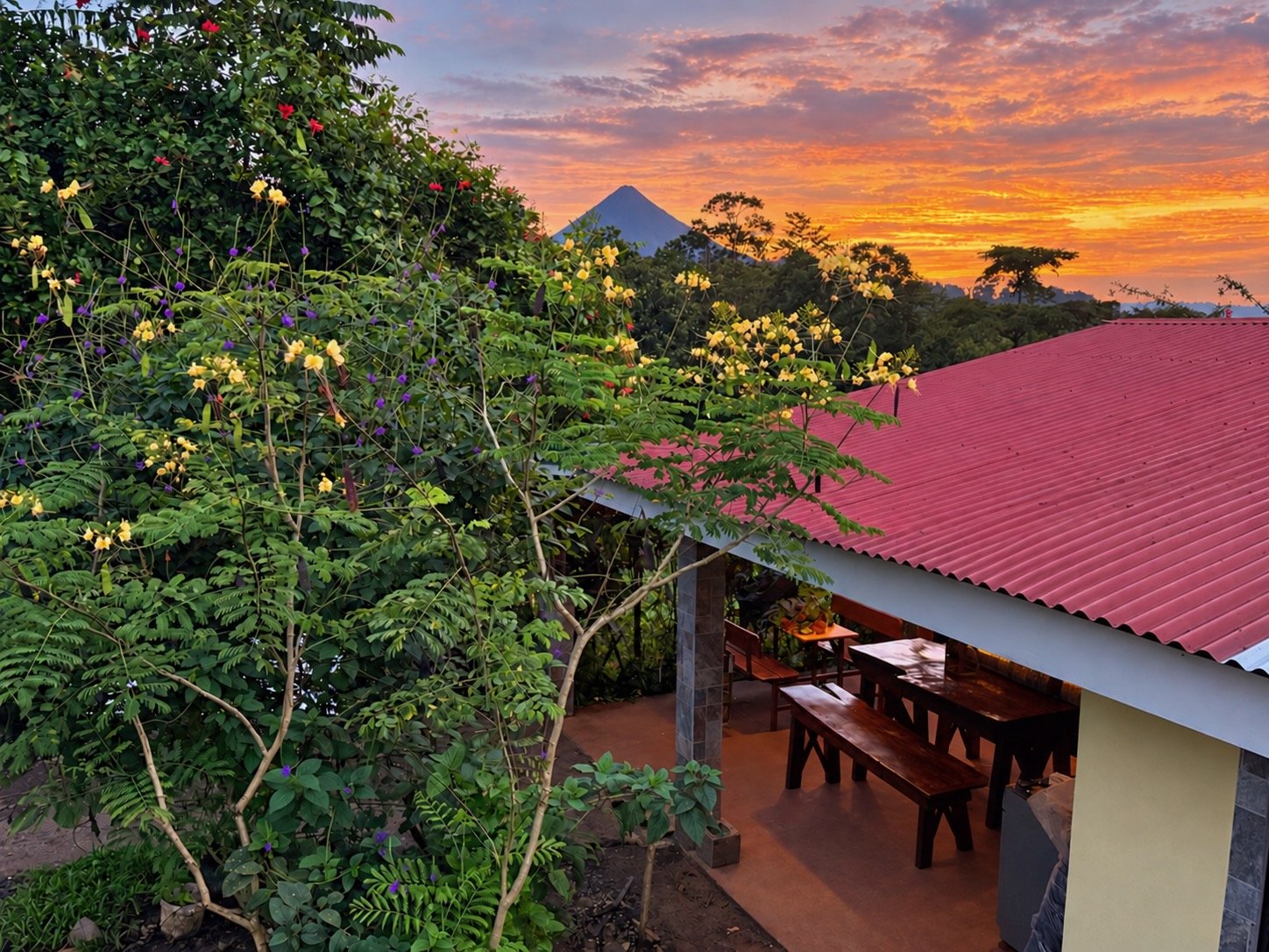 Casita Catalina patio with red corrugated roof and wooden dining benches looking out toward Arenal Volcano at sunset