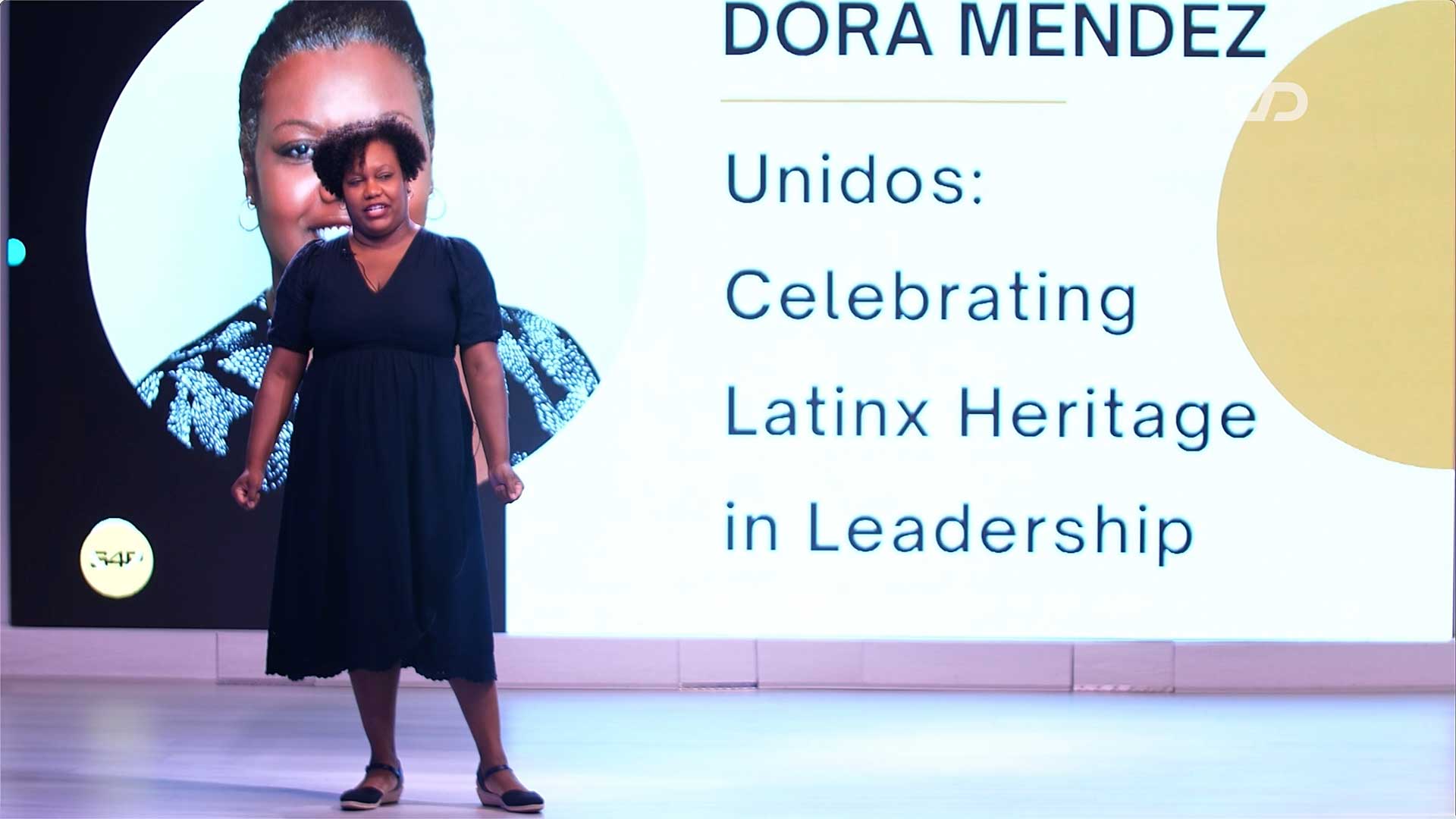 Coach Dora Mendez delivers a keynote on stage in front of a large presentation screen displaying her name and the title "Unidos: Celebrating Latinx Heritage in Leadership."