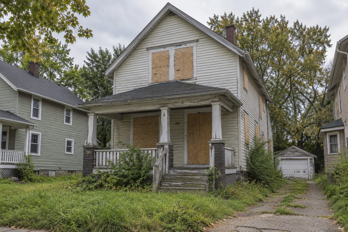 Vacant older house with boarded windows, weathered siding and an overgrown yard