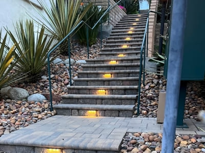 Evening view of a stairway with integrated lighting on each step, bordered by decorative rocks and desert plants, guiding safe passage up the stairs.