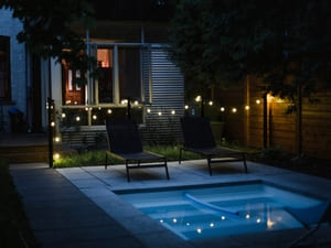 String lights and soft pool lighting create a relaxing atmosphere in a modern backyard with lounge chairs by a small pool.