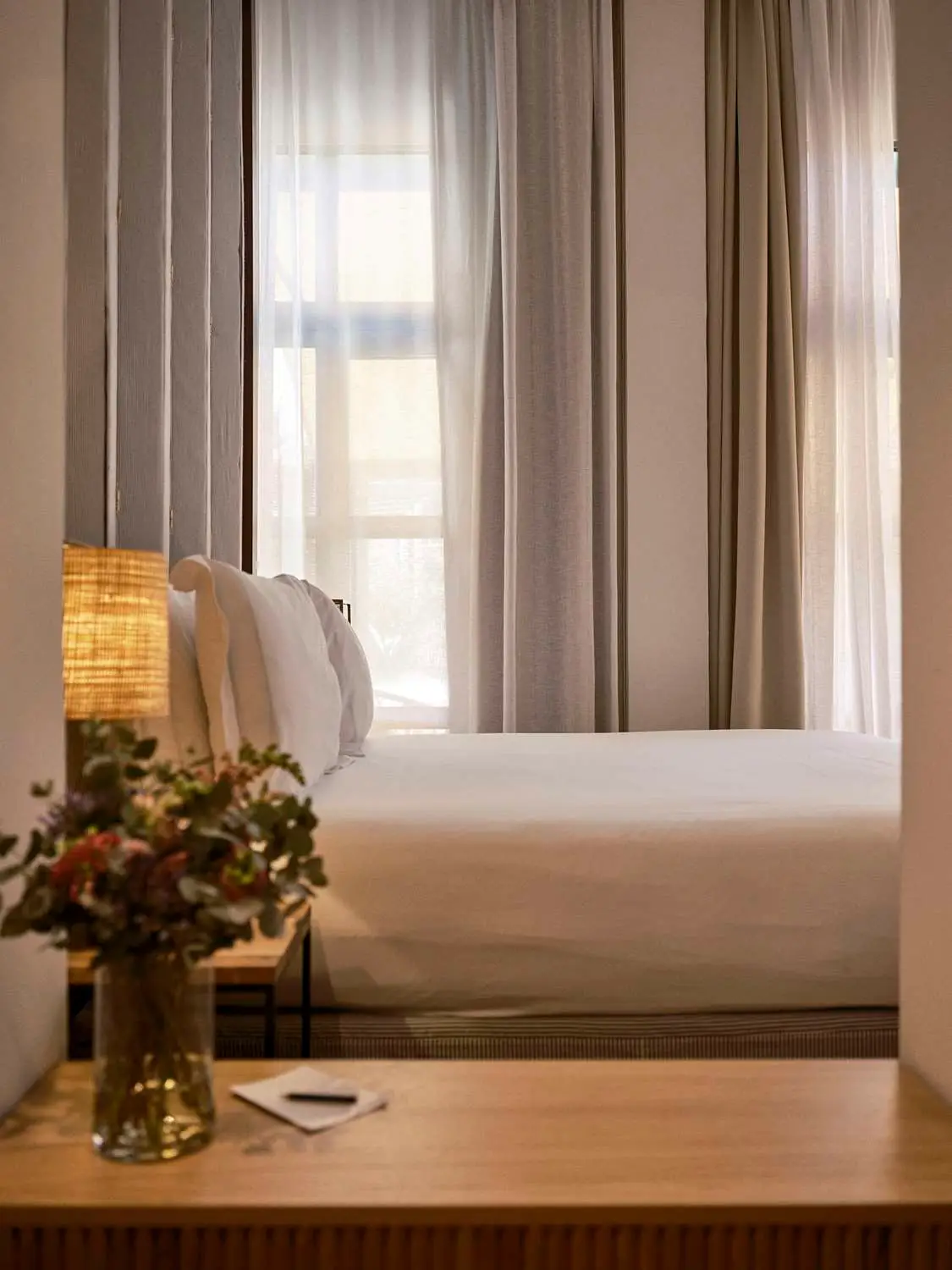 Hotel room with a made bed, soft curtains and bedside flowers Hotel room with a made bed, soft curtains and bedside flowers