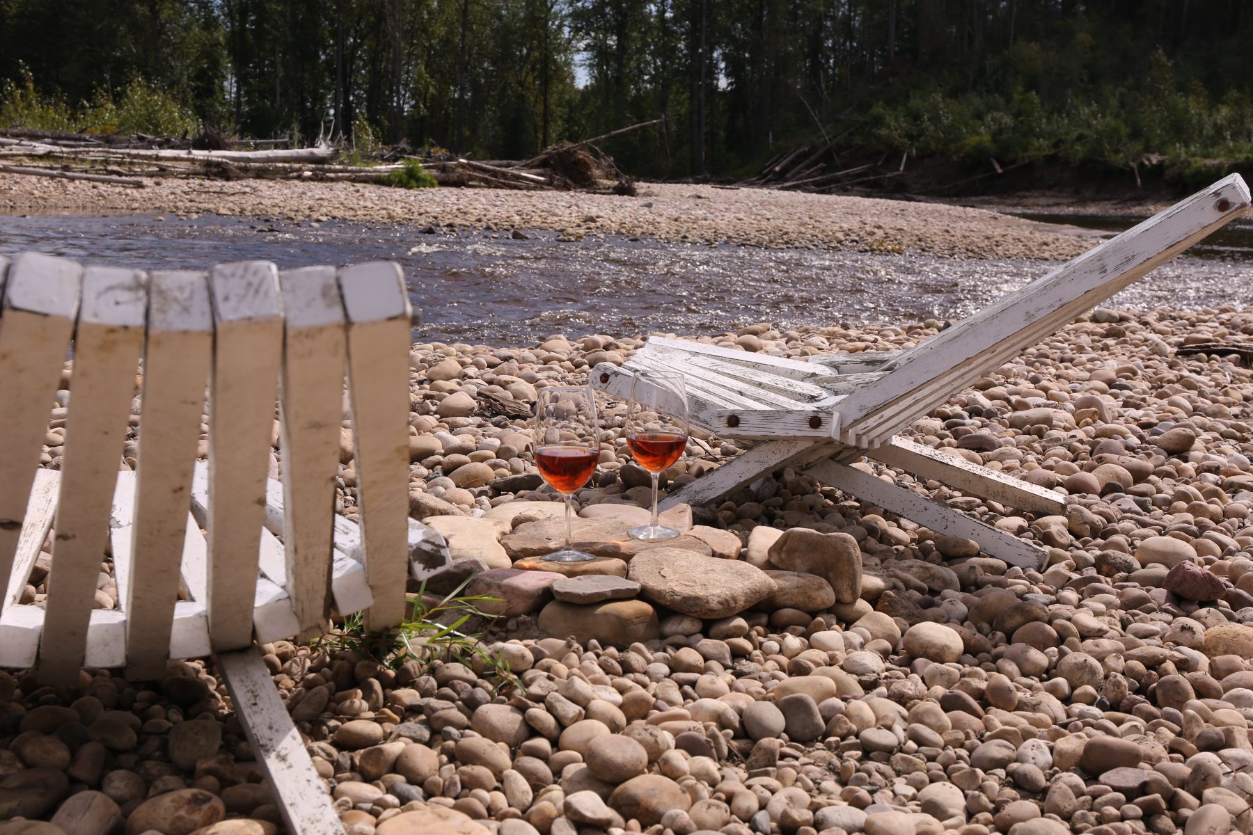 Two glasses of wine on the pebble shore beside driftwood chairs
