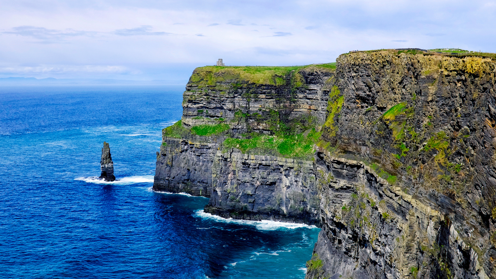 A closer view of the layered cliff face and green cliff top above blue Atlantic water