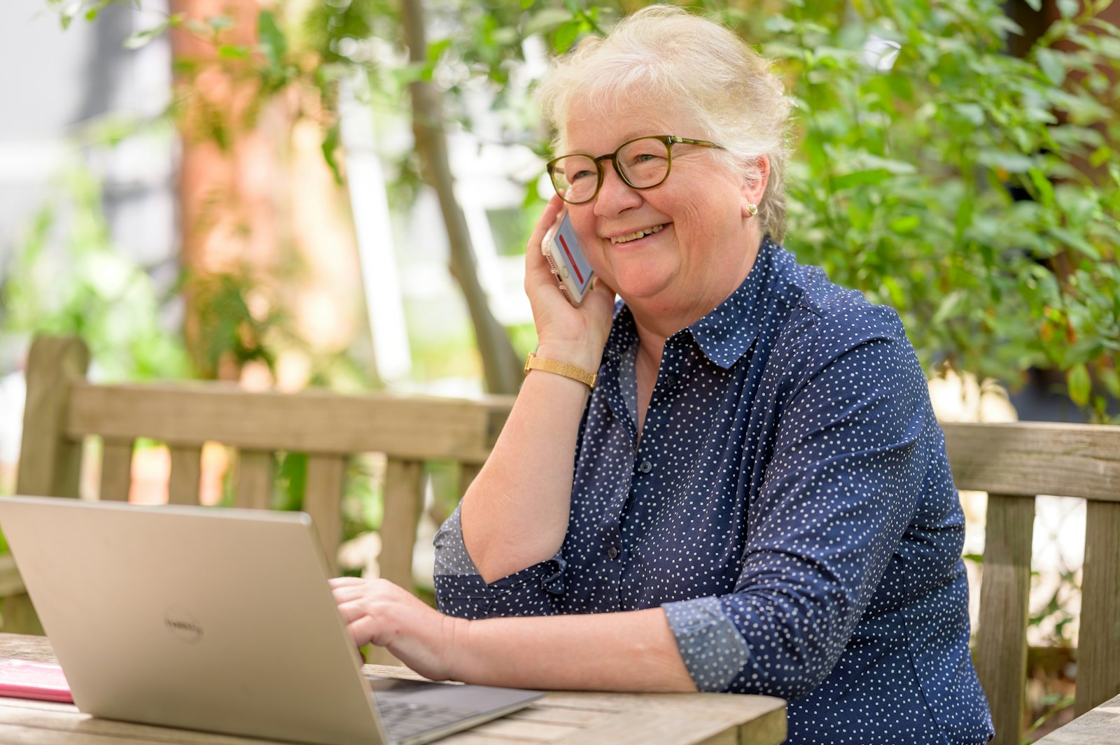 Older woman talking on the phone at an outdoor table with a laptop