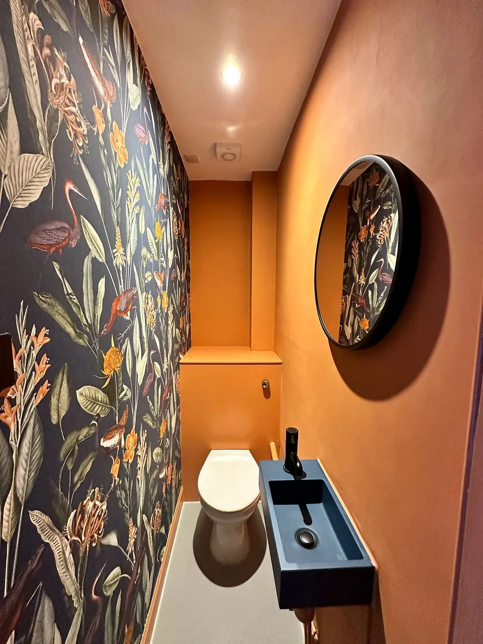 A cloakroom at Broughton Street Lane with dark floral wallpaper and a round mirror.