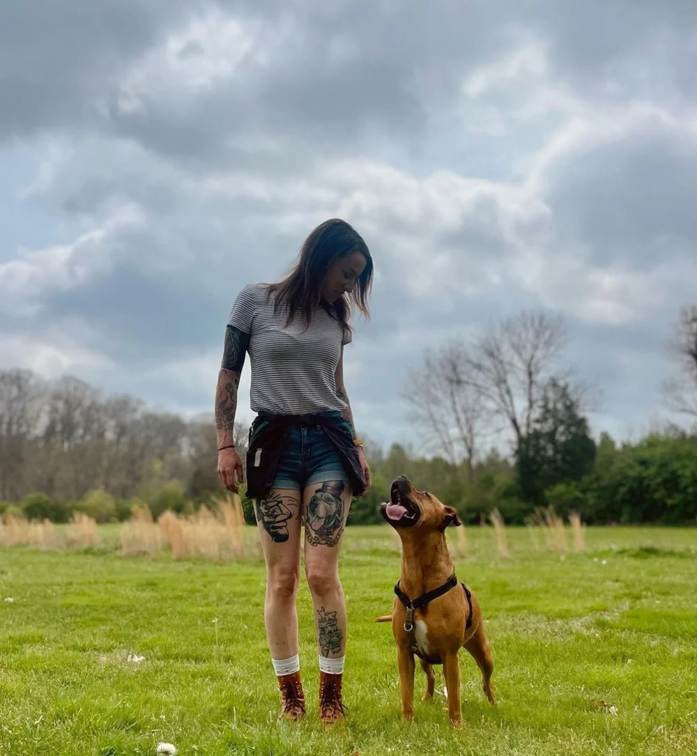 Jenna kneeling on grass outdoors, working with an adolescent dog on a loose-leash walk