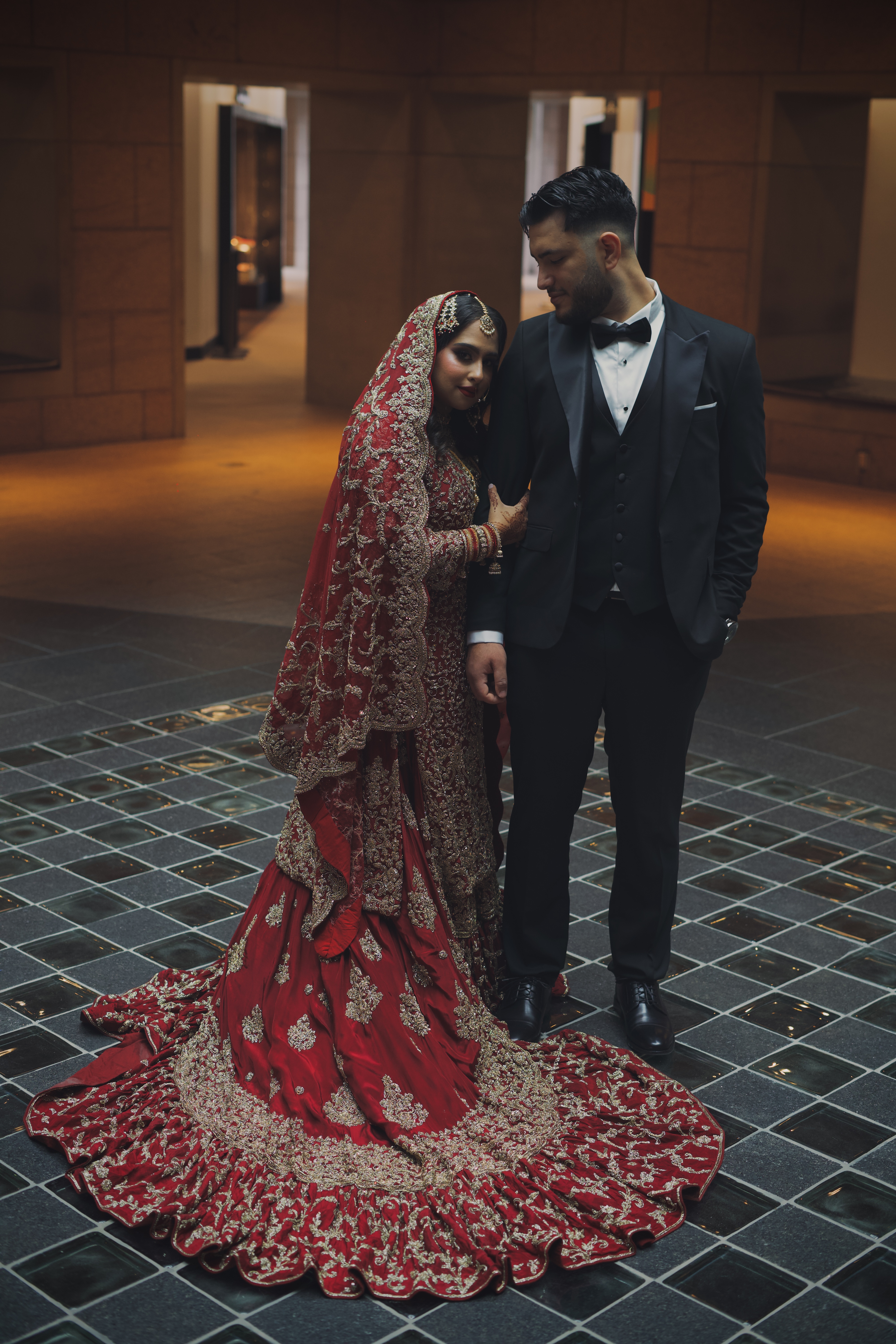 Bride in a red and gold bridal lehenga with a long train beside her tuxedoed groom in an elegant venue.