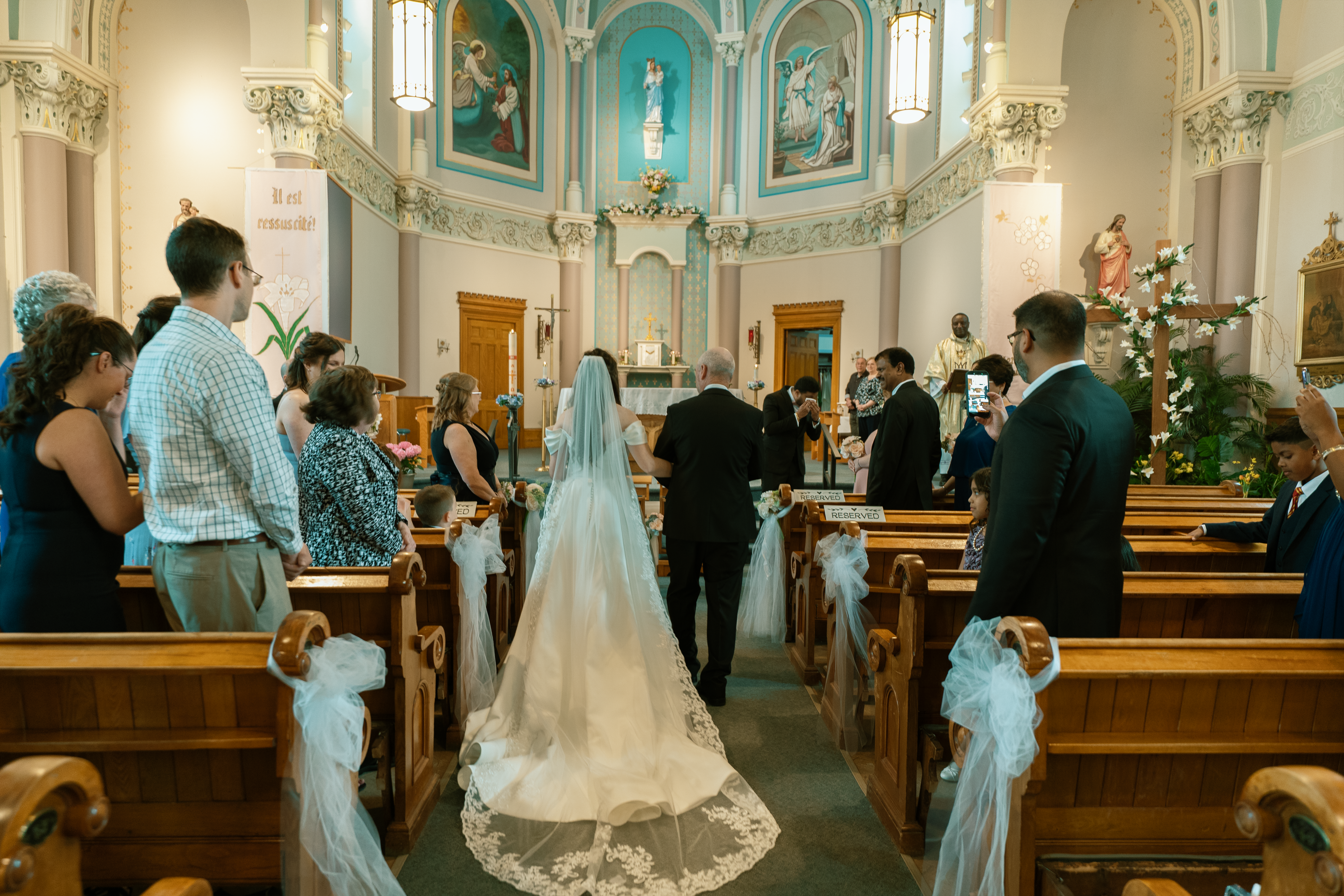 Bride in a long cathedral veil walking down the church aisle during the ceremony.