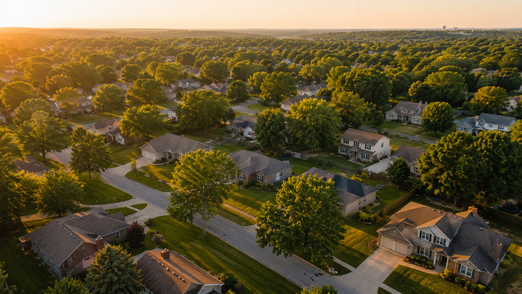 Aerial view of a tree-lined Kansas City suburban neighborhood with ranch and two-story homes illuminated by warm golden-hour sunlight.