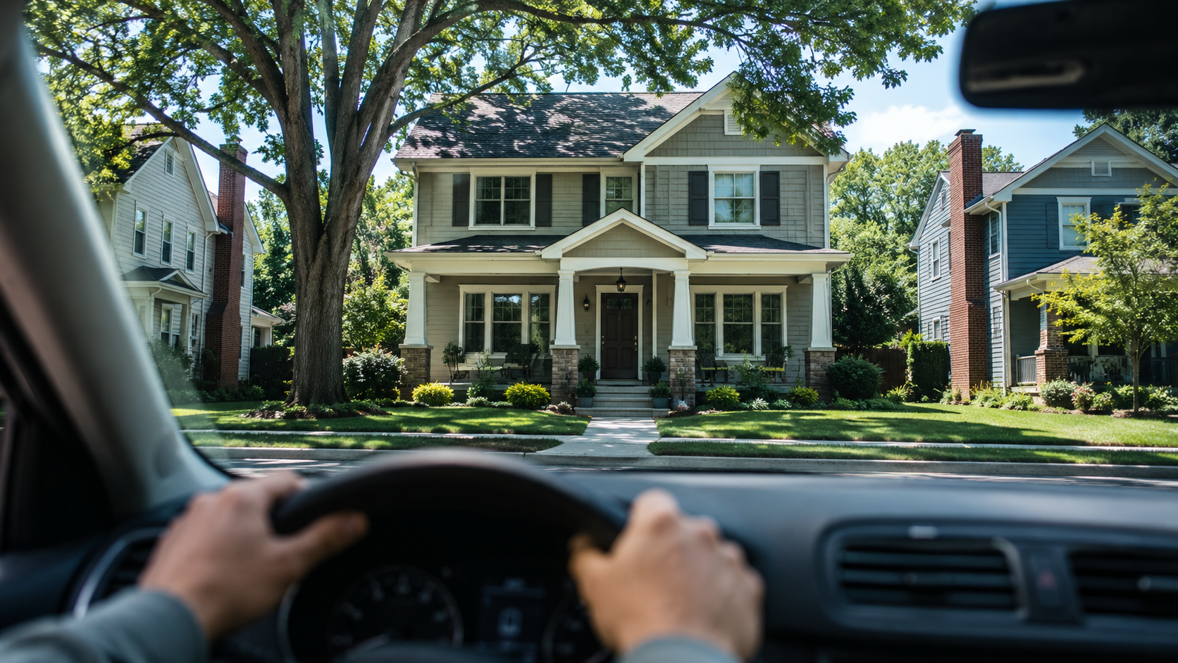 Driver’s view through a parked car windshield toward a well-kept two-story Kansas City home on a quiet, tree-lined suburban street.