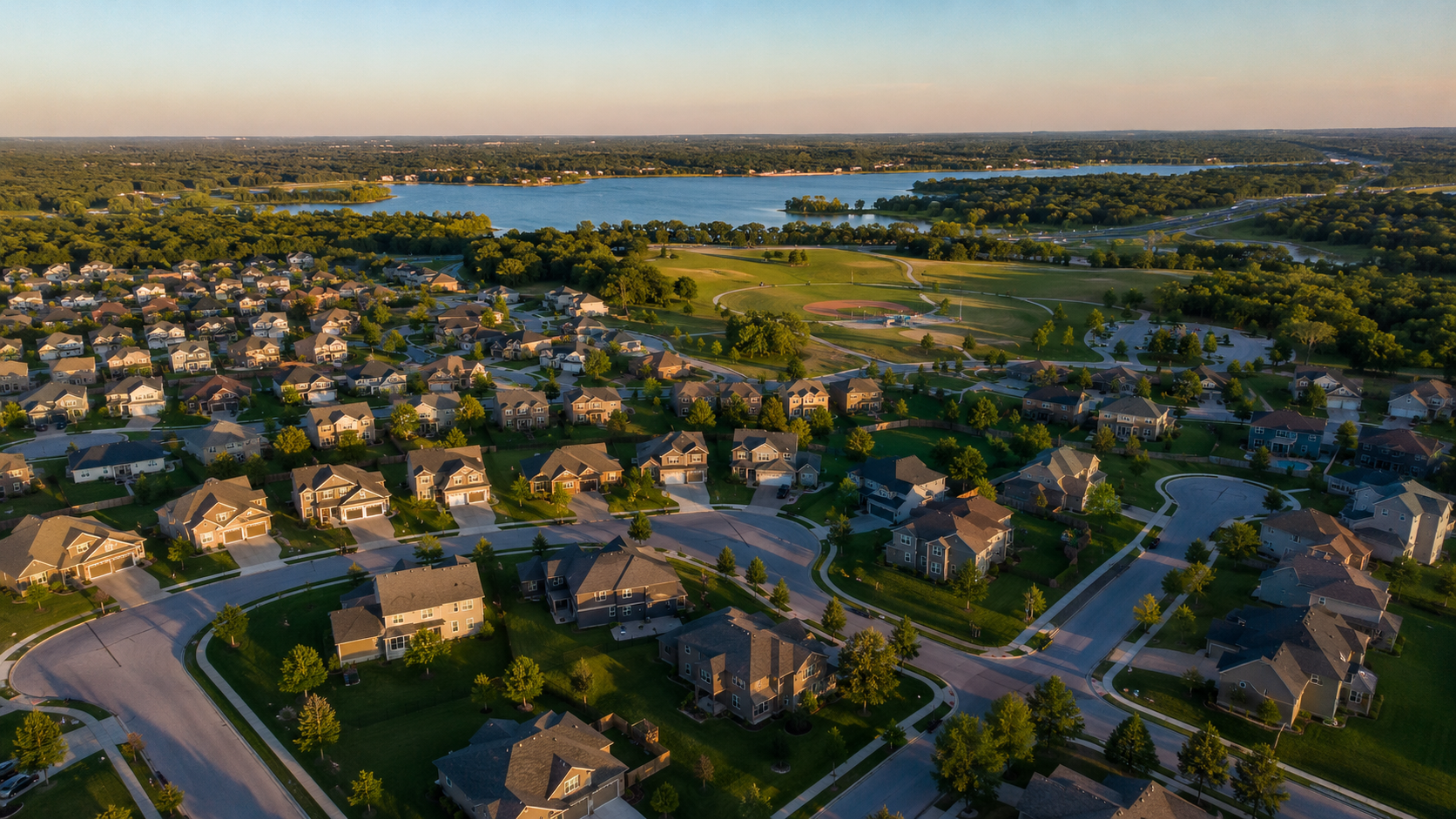Aerial view of a newer Blue Springs, Missouri, neighborhood with single-family homes, green lawns, cul-de-sacs, a large park, lake, and distant highway. Aerial view of a newer Blue Springs, Missouri, neighborhood with single-family homes, green lawns, cul-de-sacs, a large park, lake, and distant highway.
