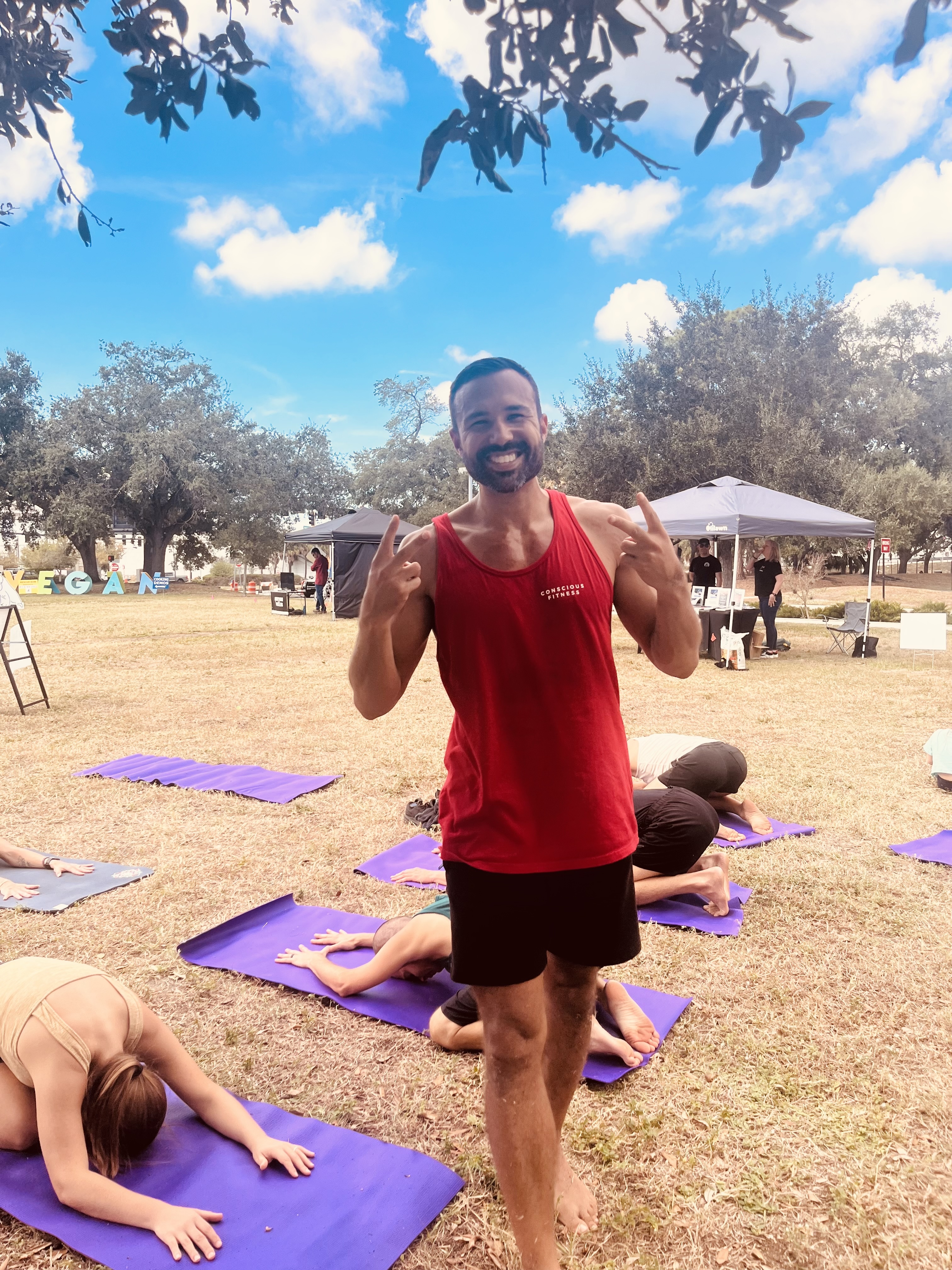Bryan smiling as he leads an outdoor strength and mobility class, participants stretching on mats behind him