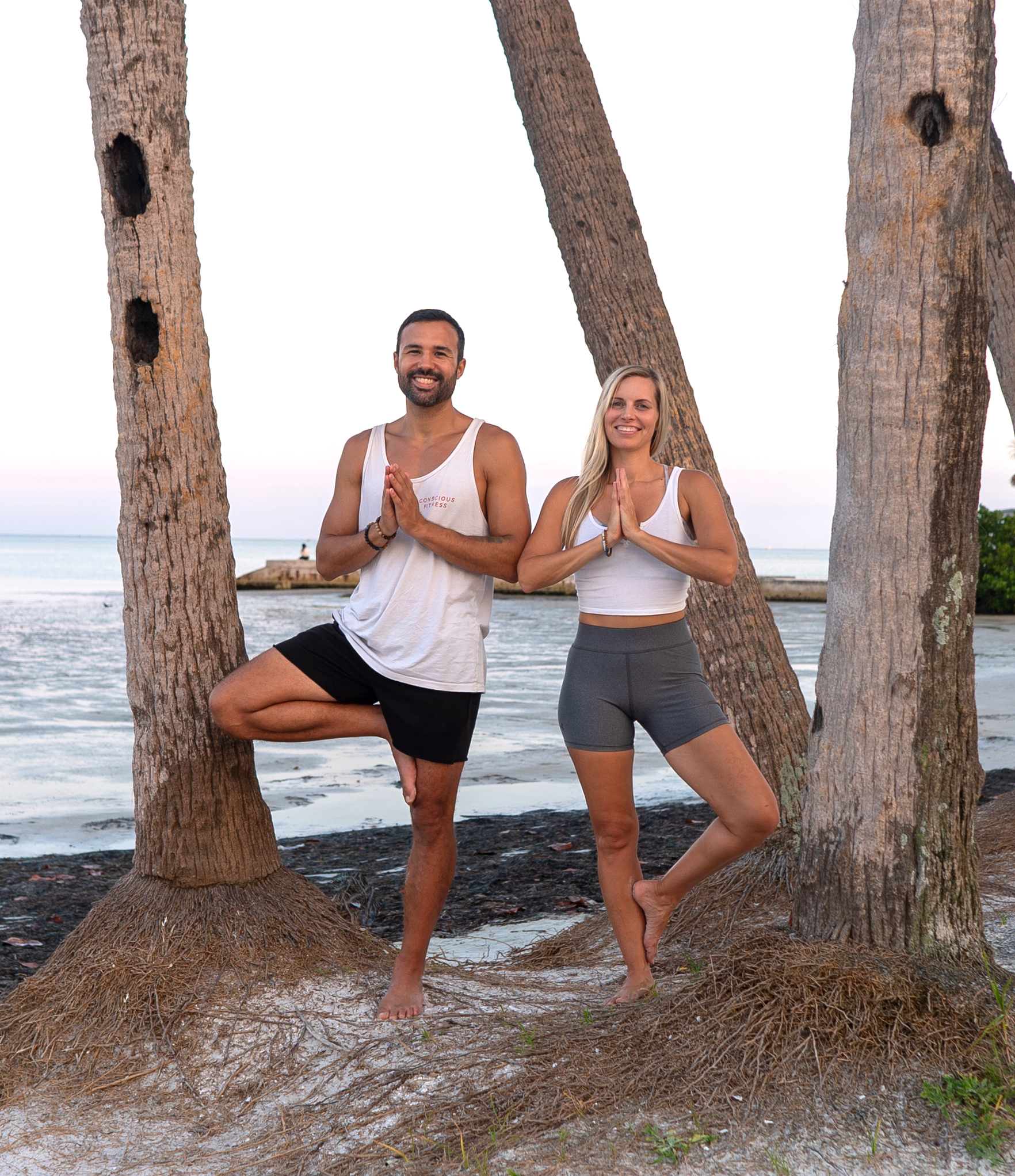 Bryan Bell and Jenny Roberts smiling in tree pose between palm trees on the beach