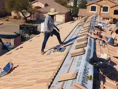 Roofer in a wide-brim hat on a tile roof, new underlayment rolled out beside him