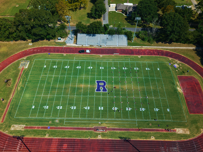 Aerial view of a high school football stadium with a marching band practicing on the field.