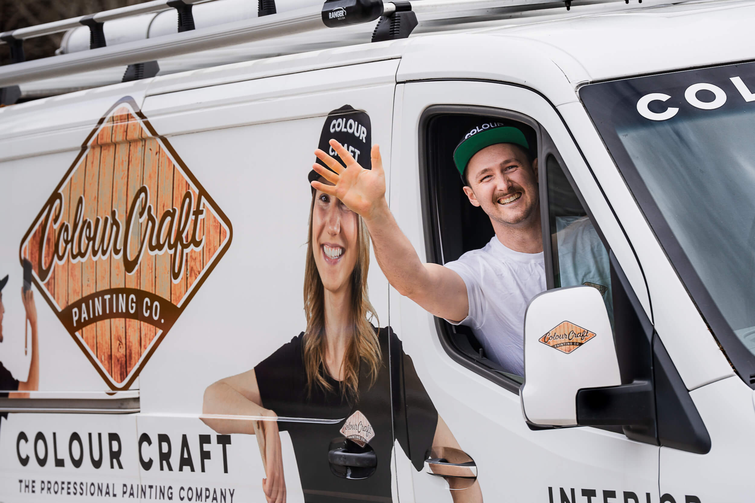 Cheerful male painter from Colour Craft waving from the driver's seat of the company's branded vehicle, with the playful graphic wrap featuring a female colleague. The van prominently displays the Colour Craft logo, emphasizing the brand's professional painting services.