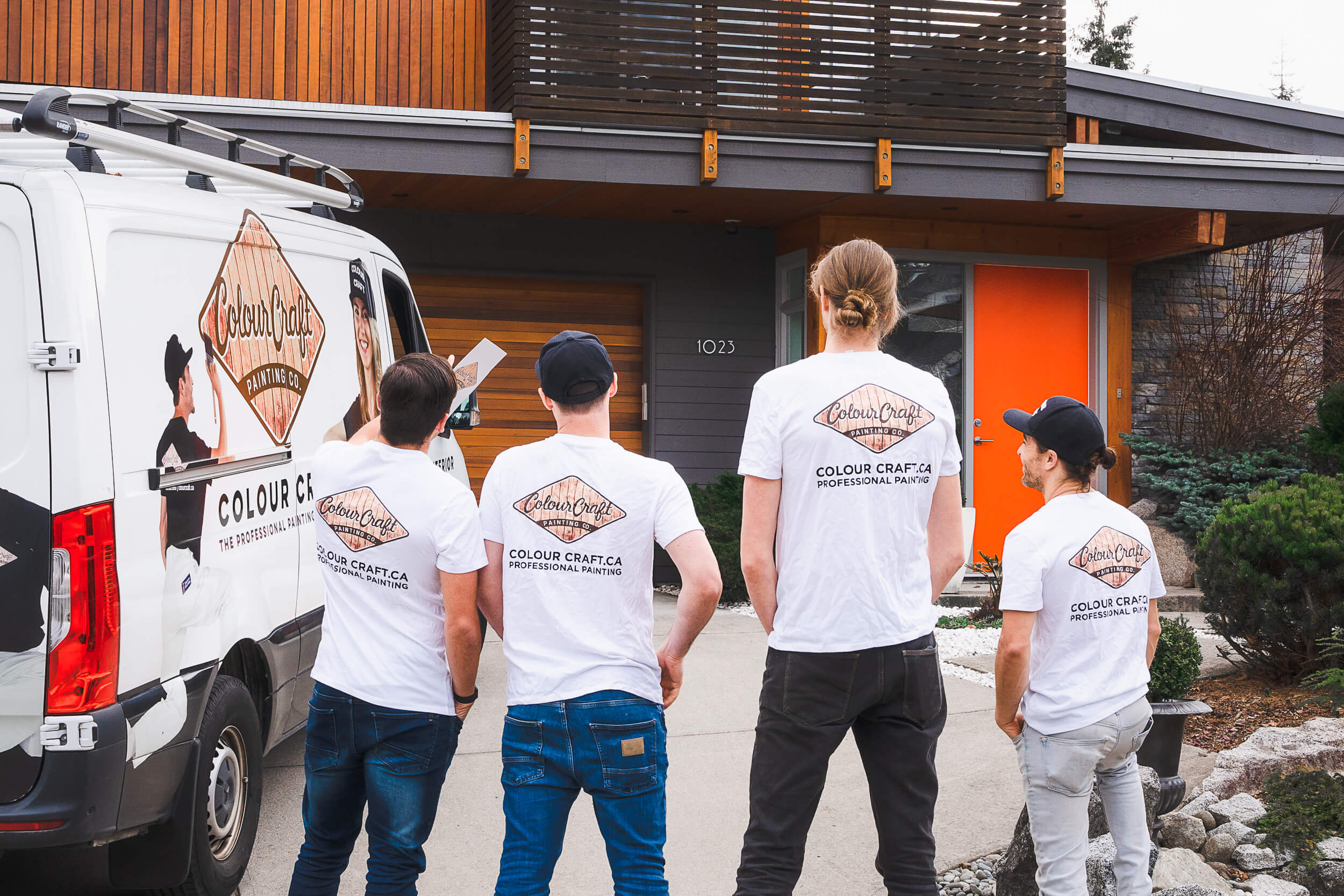 A team of friendly Colour Craft painters engaging in a lively discussion outside their service van. They are dressed in uniform white t-shirts with the company's logo, sharing a laugh while reviewing a project plan, conveying a professional yet approachable image of the company's workforce.