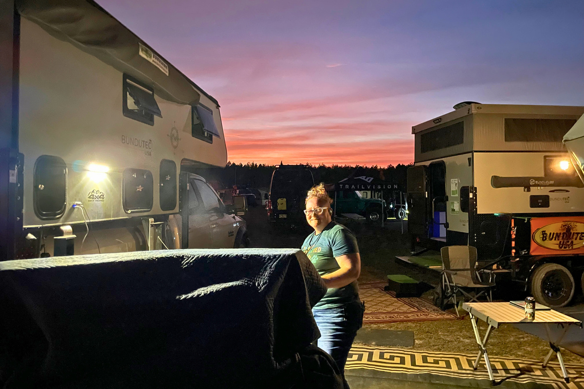 BundutecUSA truck camper display illuminated at night during the 2026 S.A.V.E. Expo, with Jenn cooking dinner on the Tembo Tusk camp stove in the foreground under a colorful dusk sky.
