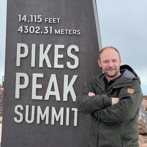 Baz Porter at Pikes Peak Summit, standing next to a sign marking 14,115 feet elevation. Leadership coach and speaker embracing personal growth, resilience, and achievement at the mountain’s peak.